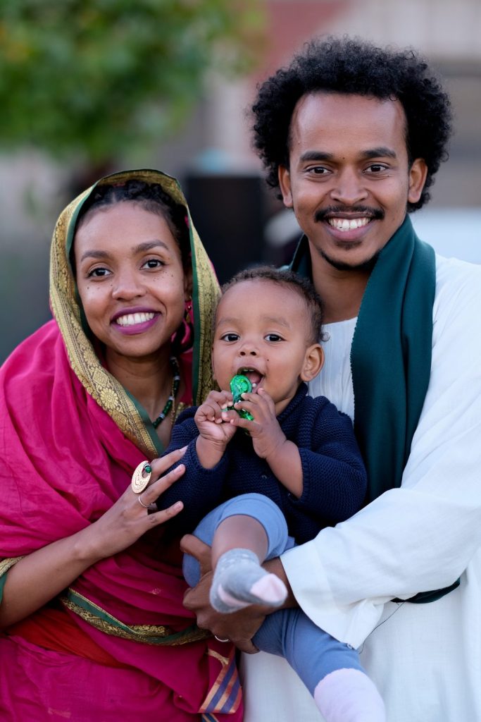 Fatima Shulli and Abdo Habbani standing outdoors, smiling, holding their baby between them.