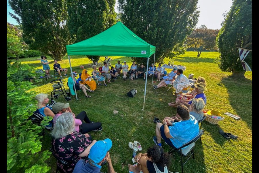 Neighbours seated in a wide circle of chairs under a green tent between trees at Churchill Park.